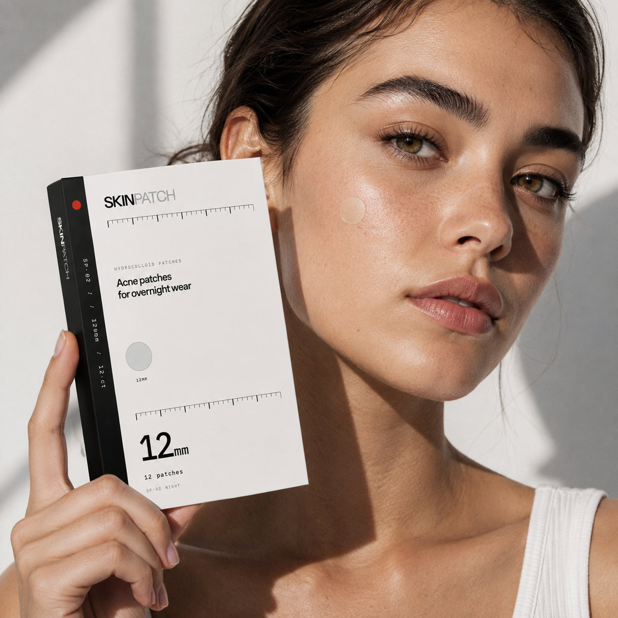 Model holding a SKINPATCH Night box, a hydrocolloid patch sealed on her cheek in raking daylight
